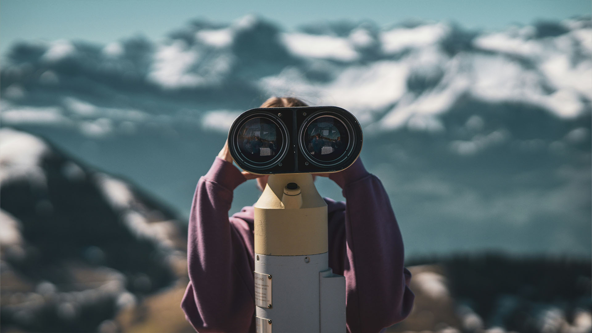 A person looking through binoculars at you, with snowy mountains in the background